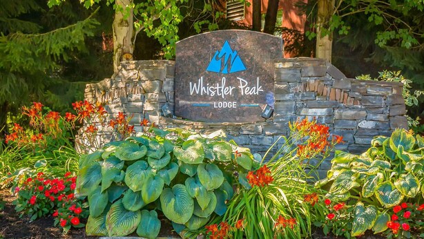 Kivestä valmistettu kyltti, jossa on logo ja teksti ”Whistler Peak Lodge”. Kylttiä ympäröi runsas vihreys ja kukkaloisto.