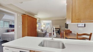 Cocina compacta con lavamanos, armarios de madera, comedor con sillas y zona de estar con sofá y televisor.