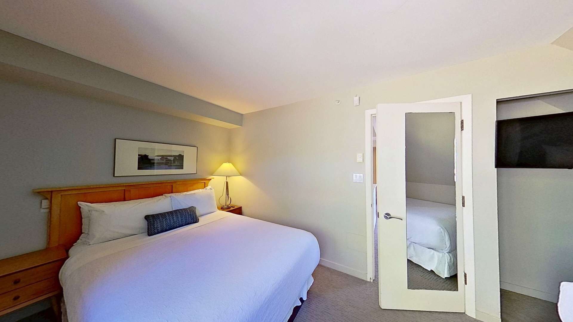 Une chambre d’hôtel avec un grand lit, une table de chevet avec une lampe, une porte donnant sur un placard et un tableau encadré au mur.