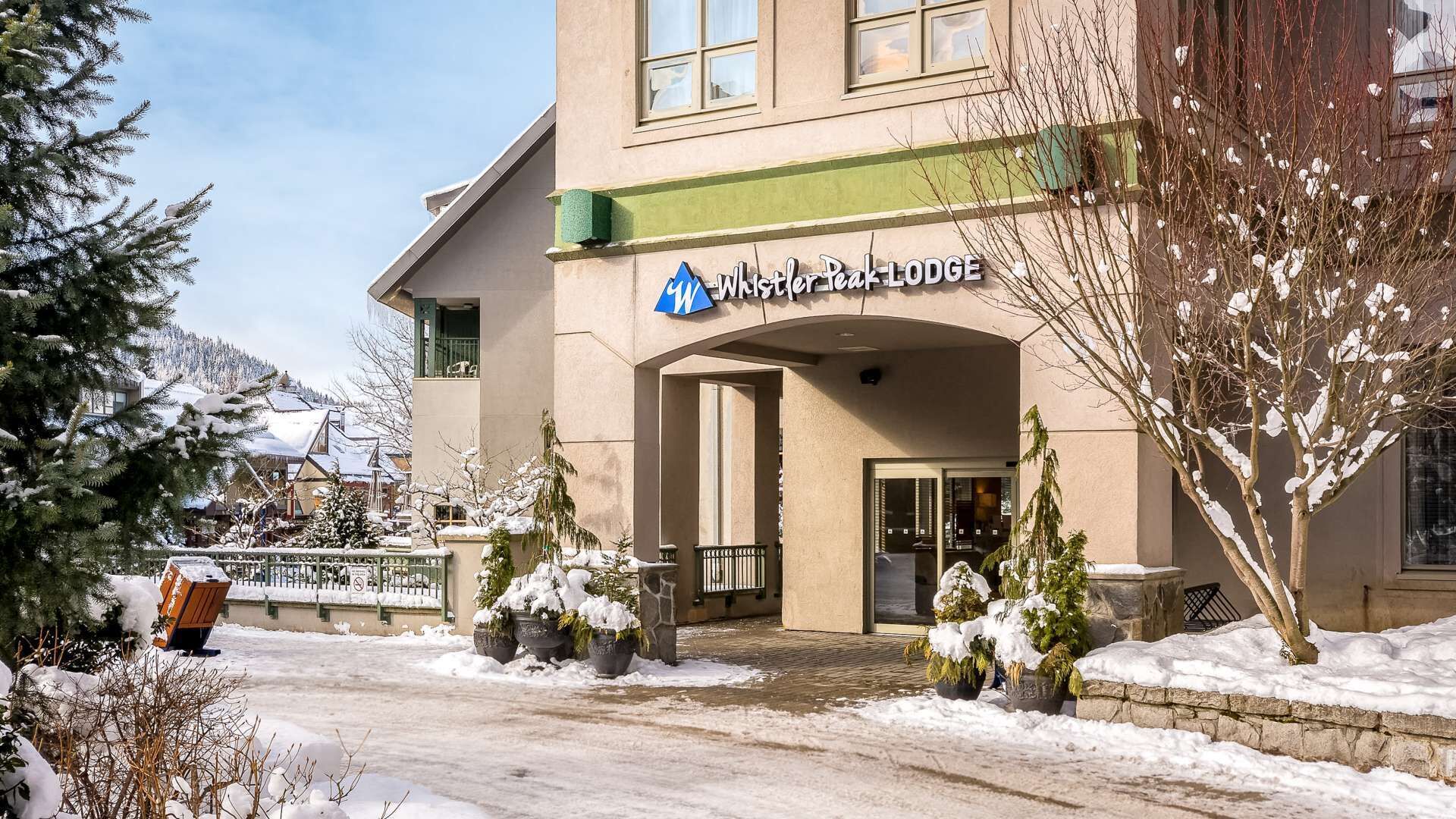 A building with a sign reading 'Whistler Peak Lodge' with a snow-covered entrance and potted plants.