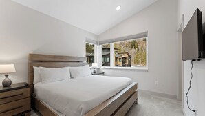 Een slaapkamer met een houten bed, een nachtkastje met een lamp, een aan de muur bevestigde televisie en een raam met uitzicht op huizen.