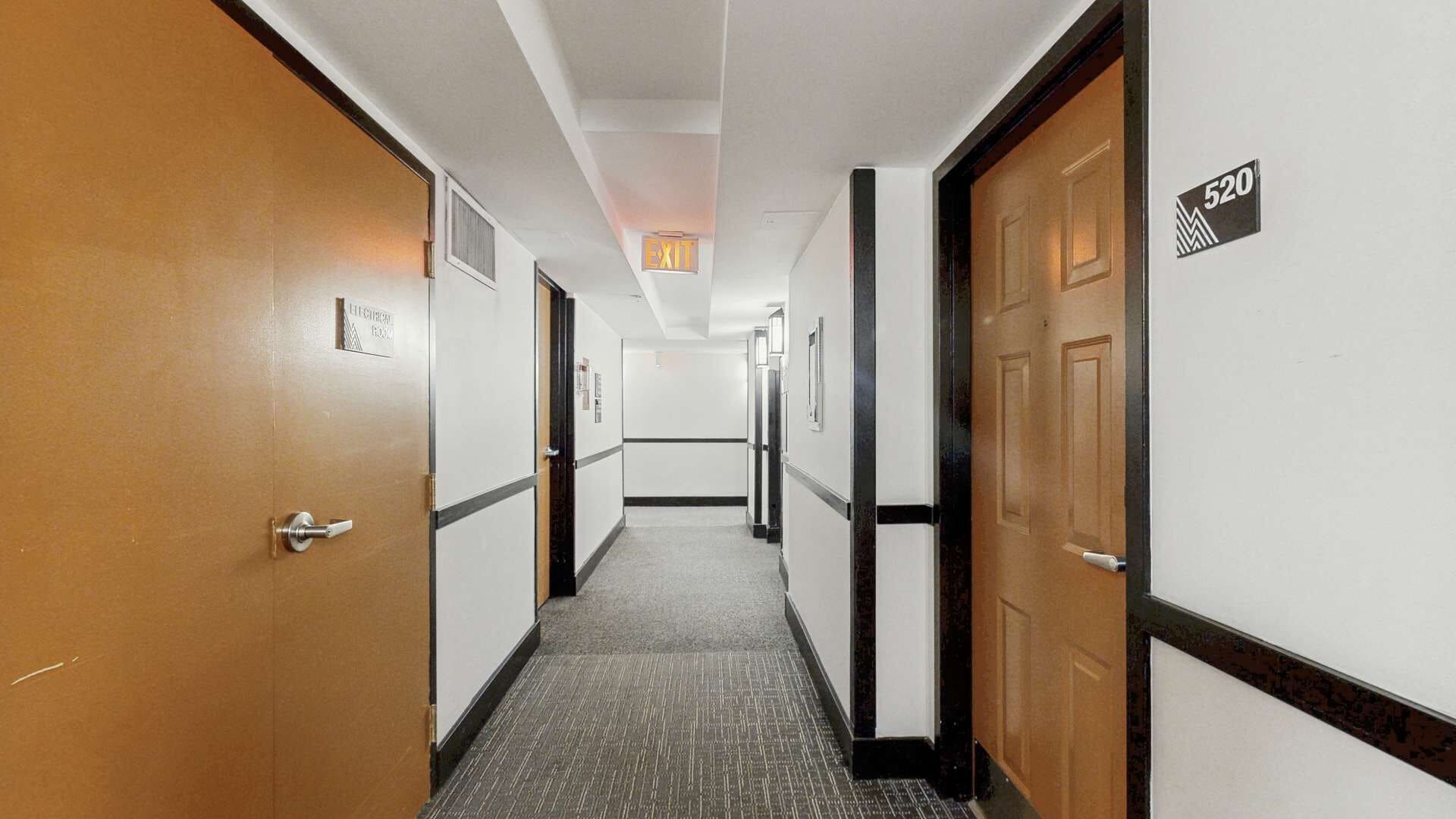 A hotel hallway with doors, a carpeted floor, and an exit sign.