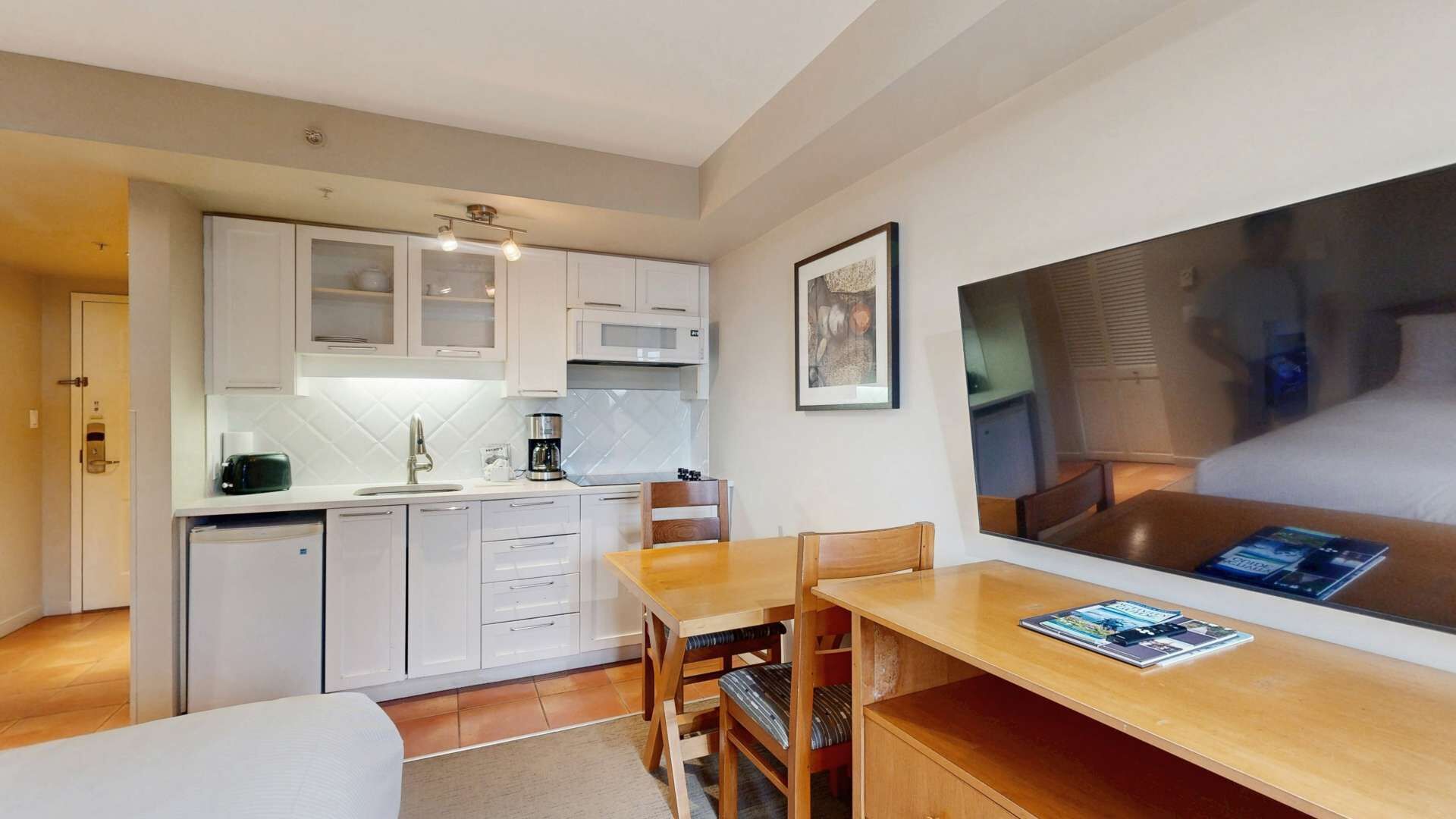 A compact kitchen with white cabinets, a microwave, and a coffee maker. There is a dining area with a wooden table and chairs, and a large framed picture on the wall.