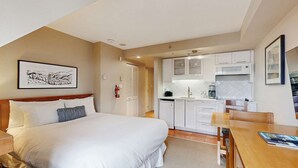 Un dormitorio con cama, mesita de noche y un pequeño escritorio con silla. Incluye una kitchenette con microondas, tostadora y cafetera. Encima de la cama hay un cuadro enmarcado.