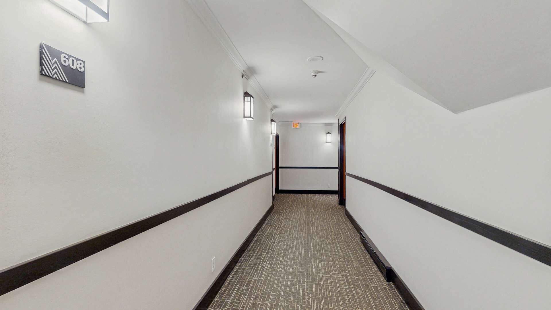 Un couloir doté d’un sol recouvert de moquette, de murs blancs et d’une bande noire qui court le long du milieu. Une enseigne portant le numéro 608 est visible sur le mur de gauche.