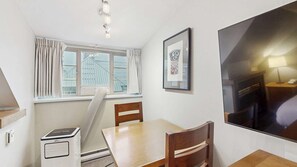 Ein Zimmer mit Esstisch, Stühlen, einem fenster mit Vorhängen, einer Heizung, einem Heizlüfter, einem gerahmten Bild und einem Fernseher.