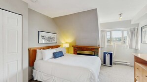 Una habitación de hotel con una cama, una mesita de noche con lámpara, un radiador, una ventana con cortinas y un cuadro en la pared.