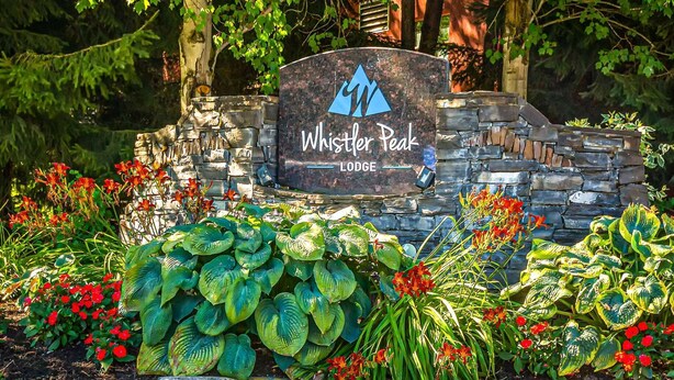 Kivestä tehty kyltti, jossa lukee ”Whistler Peak Lodge”, ja sen ympärillä on rehevää kasvillisuutta ja värikkäitä kukkia.