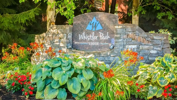 Ein Steinschild mit einem Logo und der Aufschrift 'Whistler Peak Lodge', umgeben von üppigem Grün und blühenden Pflanzen.