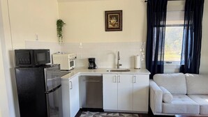 A compact kitchen with a refrigerator, microwave, coffee maker, and dishwasher. There's a window with curtains and a sofa in the adjacent living area. - Cosy Red Cottage in Private Village On VT-100 (5-min to Mount Snow) (West Dover)