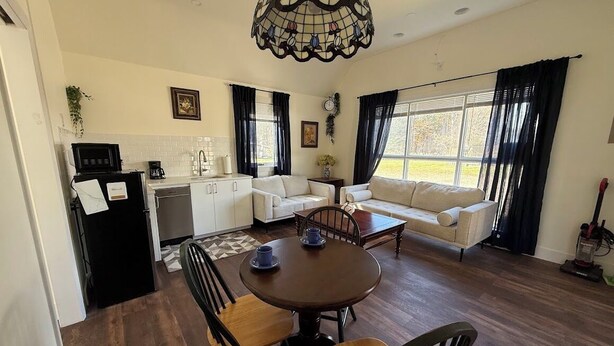 A spacious living area with a dining table, chairs, and a sofa. There is a kitchen area with a microwave and a coffee maker. The room has large windows with curtains. - Cosy Red Cottage in Private Village On VT-100 (5-min to Mount Snow) (West Dover)