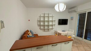Kompakti keittiö, jossa on puinen työtaso, valkoiset kaapit, ruokapöytä tuoleineen, seinälle asennettu televisio ja suuri ikkuna, jossa on sälekaihtimet.