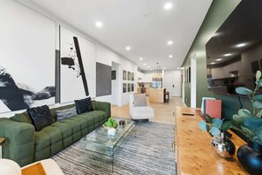 A modern living room with a green sofa, a glass coffee table, and a dining area with a wooden table and chairs.