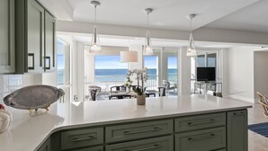 Une cuisine moderne dotée d’un grand îlot central, de luminaires suspendus et offrant une vue sur l’océan.