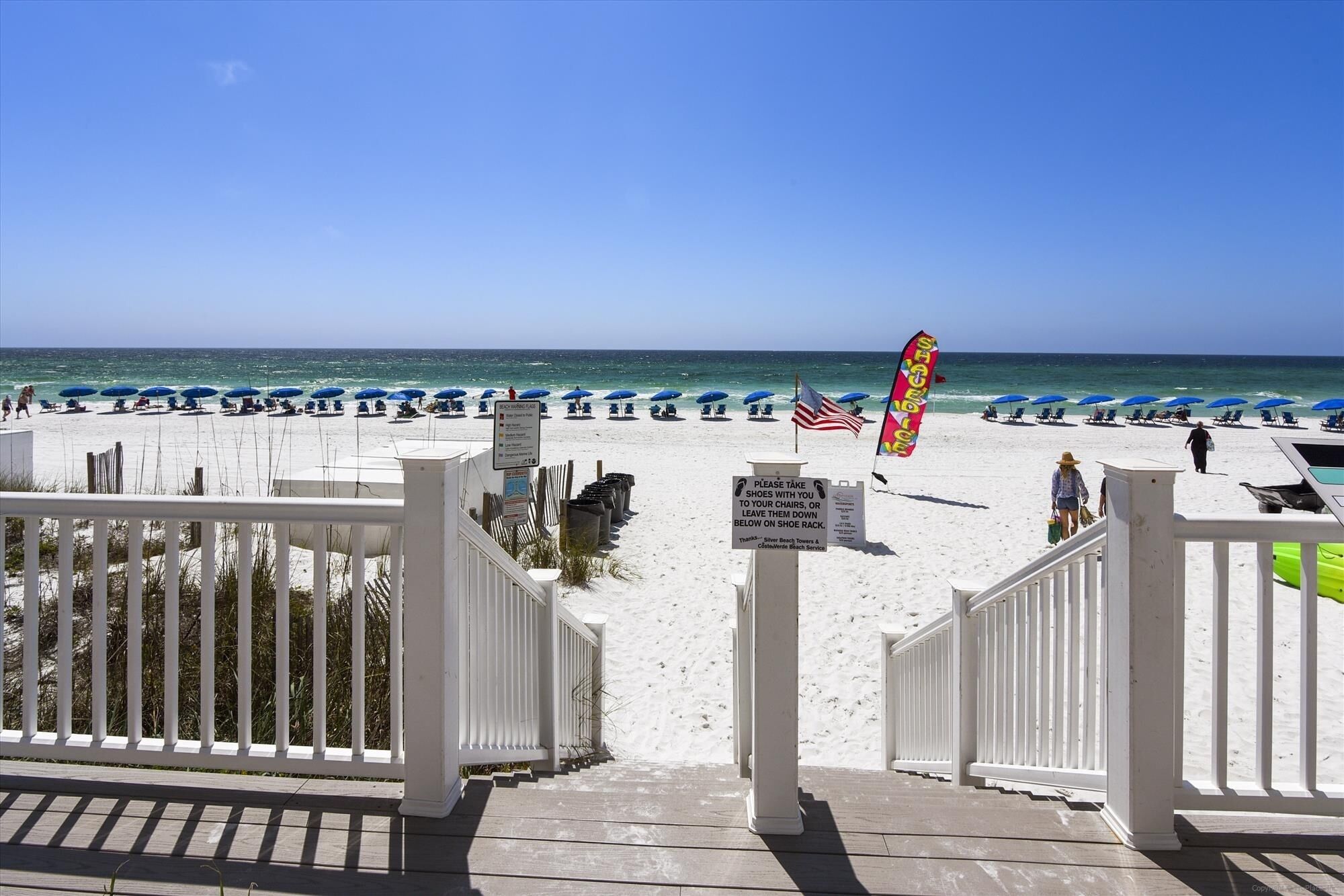 A beach view with a white fence, blue umbrellas, and a sign instructing to take shoes to chairs.