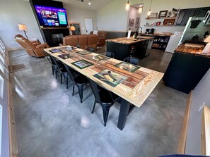 A dining area with a long wooden table, black chairs, a kitchen in the background, and a TV on the wall.