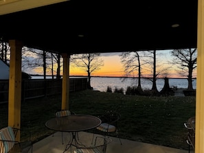 A covered patio with a view of a sunset over a body of water, a wooden fence, and a grill.
