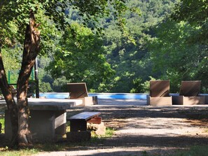 Ein ruhiger Sitzbereich im Freien mit Korbstühlen und einem Betontisch, umgeben von üppigem Grün und Blick auf ein Gewässer.