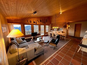 En hyggelig stue med sofa, sofabord og fjernsyn, et spiseområde med stole og bord, samt et køkken med træskabe og et vindue med udsigt til bjerge.