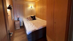 Een kleine, knusse kamer met houten wanden en een bed met een witte deken. Er staat een nachtkastje met een lamp en een klein plankje met decoratieve spullen.
