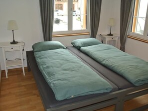 Un lit simple avec un matelas gris et une literie de couleur turquoise, flanqué de deux tables de chevet avec des lampes, une fenêtre avec des rideaux et une vue sur les voitures garées à l’extérieur.