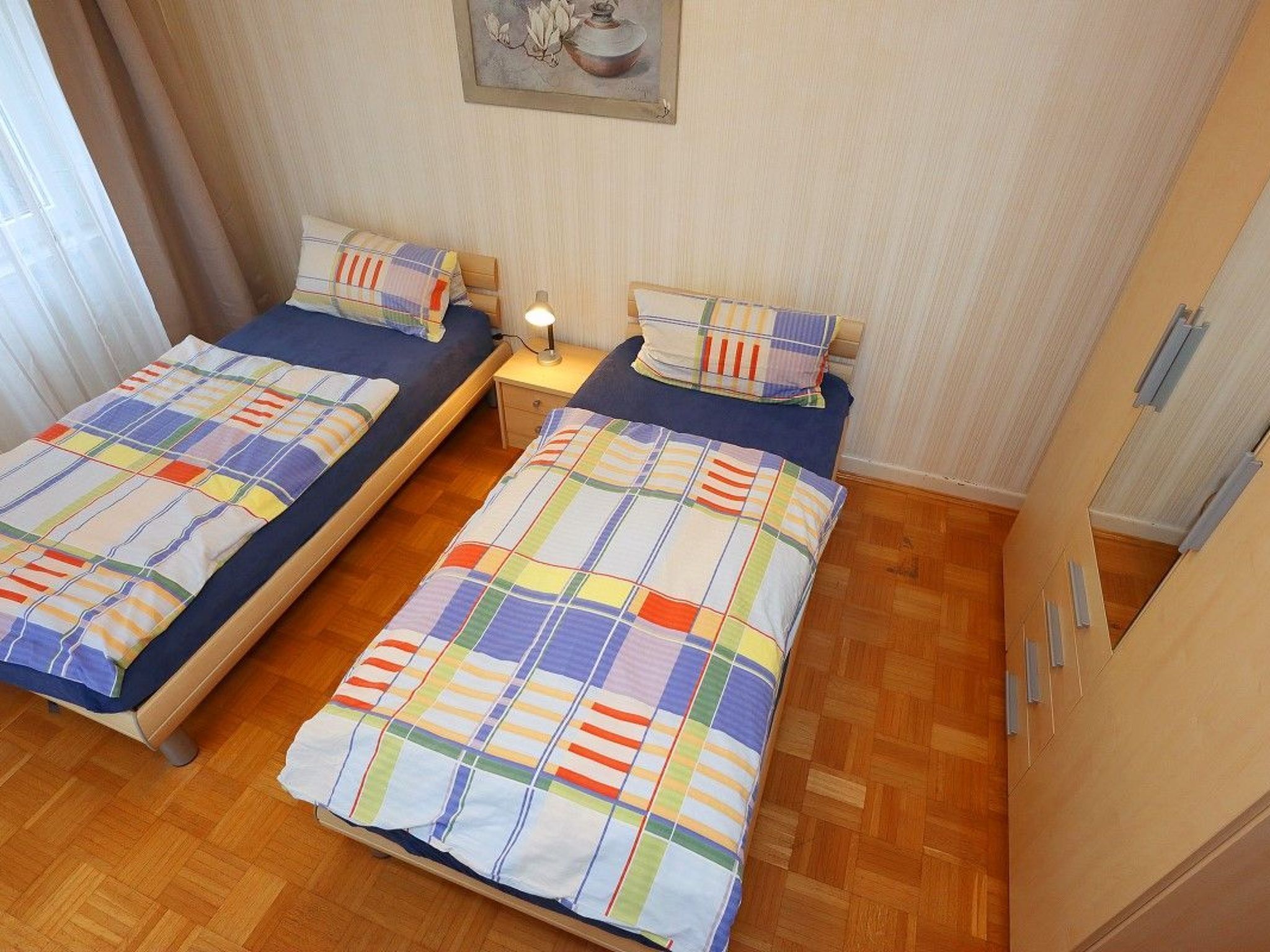 A room with two single beds, each with a colorful striped bedspread and matching pillows. There is a small bedside table with a lamp between the beds.