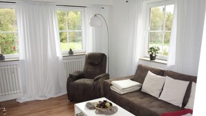 A living room with a brown sofa, a white coffee table, and a chair. There are two windows with white curtains, a radiator, and a floor lamp. - Tolle Wohnung in Klüsserath mit Großem Garten by Interhome (Klüsserath)