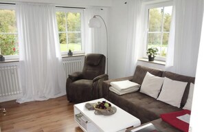 Un salon avec un canapé marron, une table basse blanche et un fauteuil. Il y a deux fenêtres avec des rideaux blancs, un radiateur et un lampadaire.