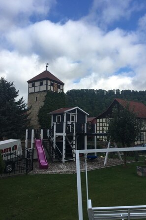 Um parquinho com escorrega e estruturas de escalada, uma torre de vigia e um prédio com telhado vermelho.