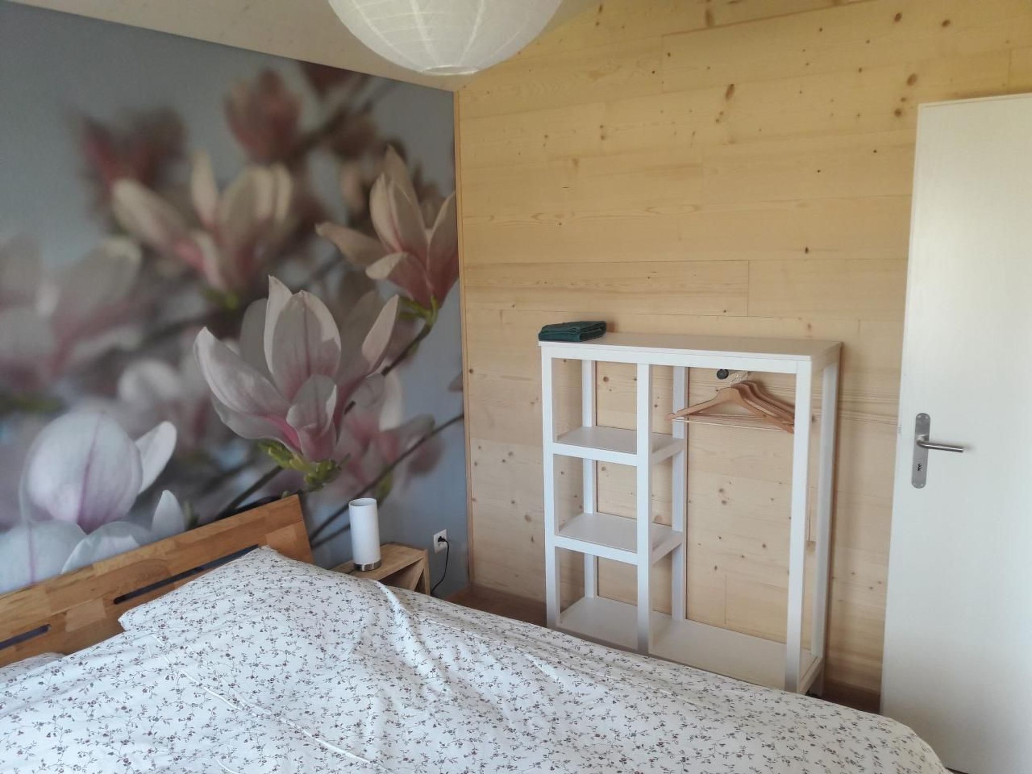 A bedroom with a bed, a wooden wardrobe, a wall-mounted shelf, and a floral mural.