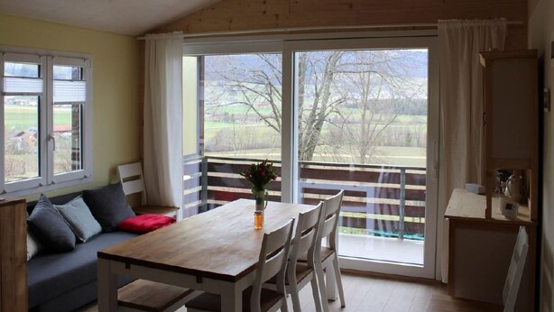 Una stanza con soffitto in legno, un tavolo da pranzo con sedie, un divano con cuscini e un'ampia finestra vista alberi e campi.