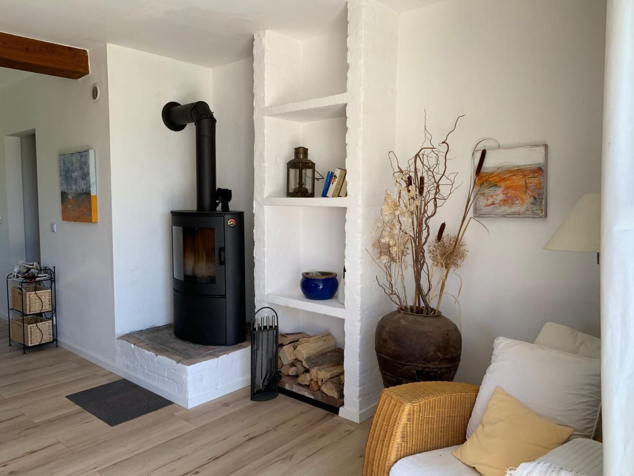 Una sala de estar acogedora con un estufa de leña negra, chimenea de ladrillo blanco, muebles de mimbre y un tazón azul sobre un estante blanco.