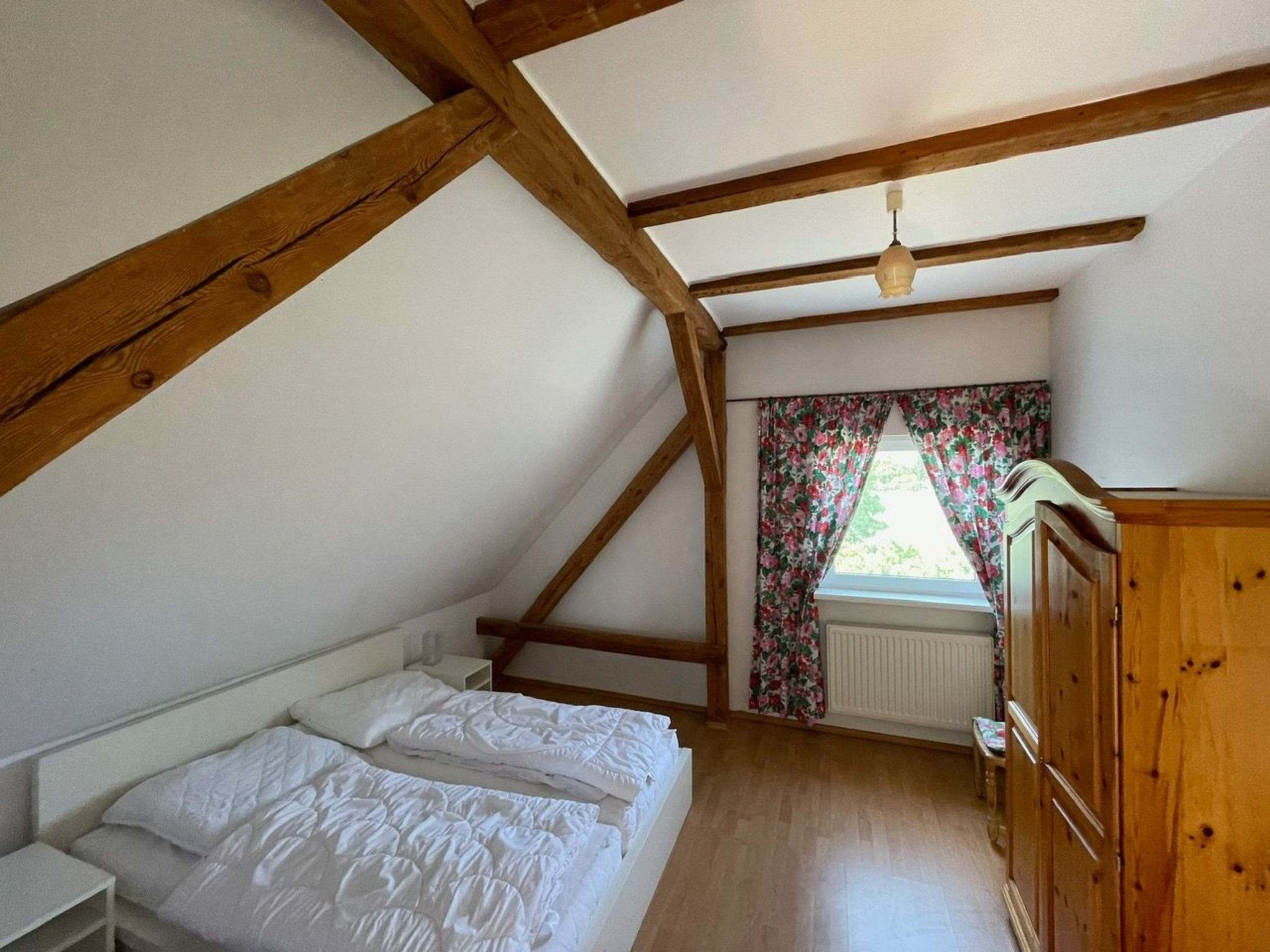 Ein Schlafzimmer mit schrägen Wänden, Holzbalken, einem Bett mit weißer Bettwäsche, einem Fenster mit blumigen Vorhängen und einem hölzernen Kleiderschrank.