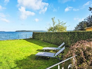 Un espace extérieur verdoyant, avec deux chaises longues donnant sur une étendue d’eau et une rive éloignée.