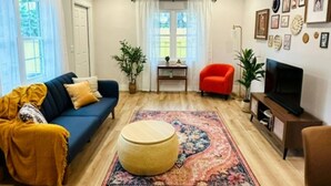 Ett vardagsrum med en blå soffa, en röd fåtölj, en rund pall, ett TV-bord och en mönstrad matta.