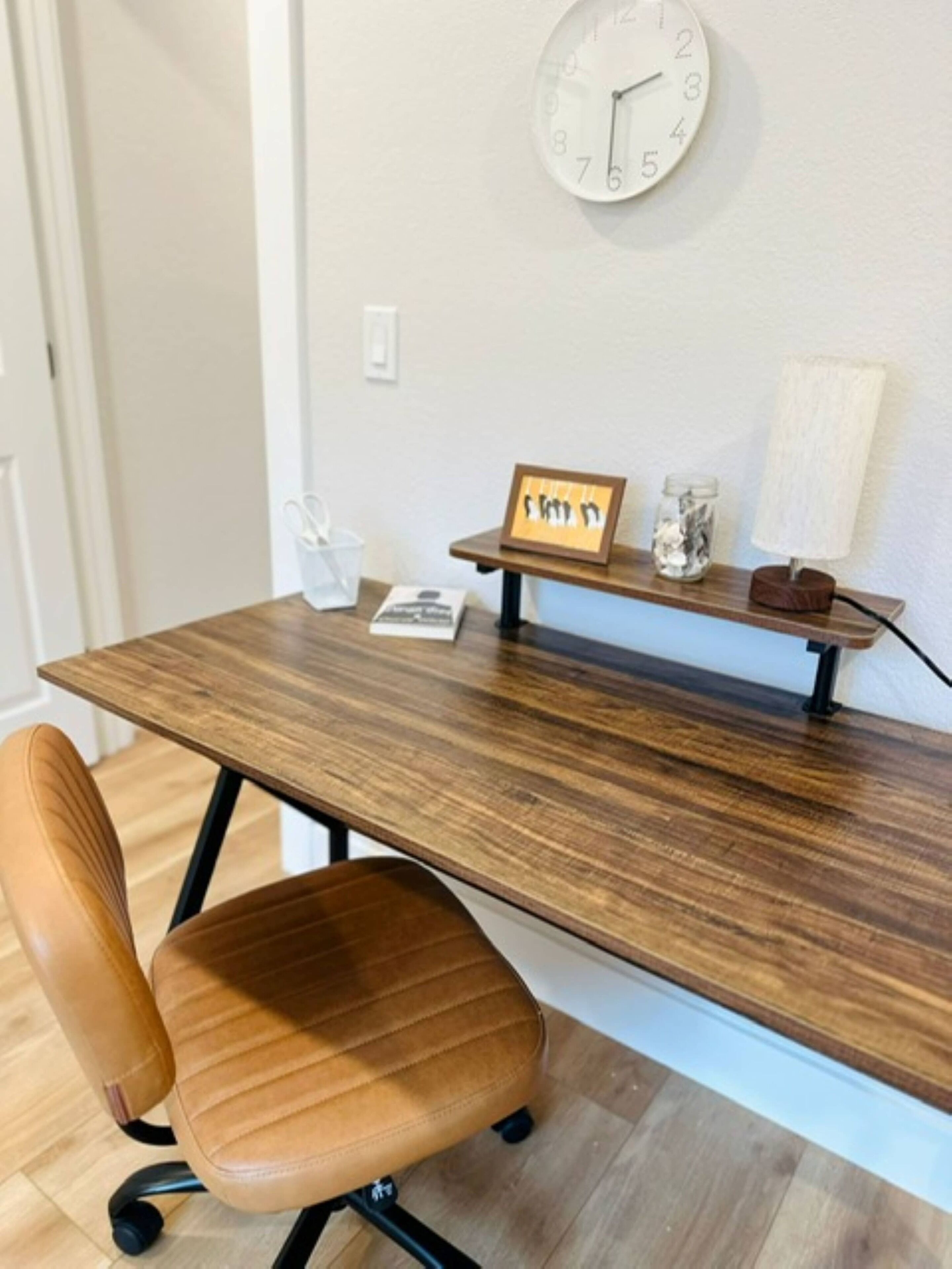 Een houten bureau met een stoel, een klok en een klein plankje met een fotolijstje en een pot.