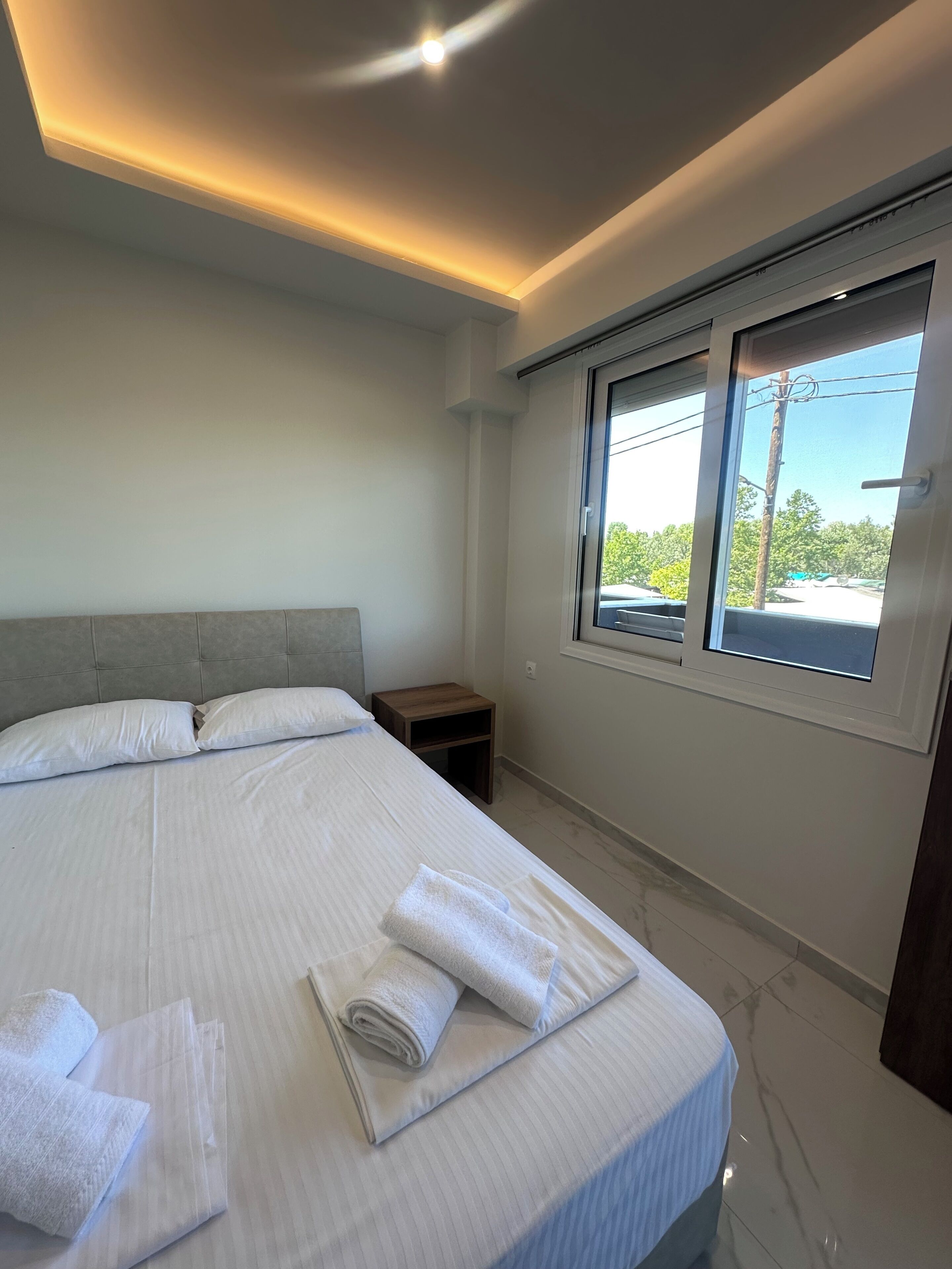 A modern hotel room with a large bed, a nightstand, a window with a view, and recessed ceiling lighting.