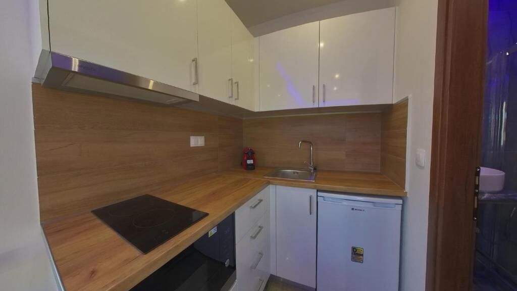 Una cucina compatta con mobili bianchi, un piano di lavoro in legno, un piano cottura integrato, un lavello e un piccolo frigorifero.