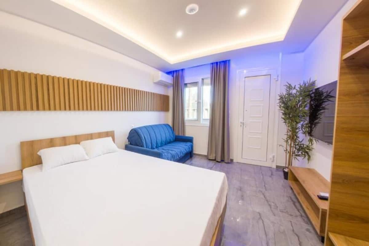Une chambre d’hôtel moderne avec un grand lit, un canapé bleu, une tête de lit en bois, un banc en bois et une plante en pot.