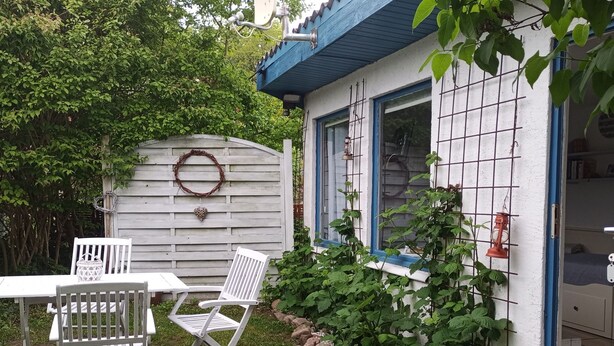 Une petite maison à l’extérieur blanc, avec des bordures bleues. On y trouve une table blanche et deux chaises blanches à l’extérieur.