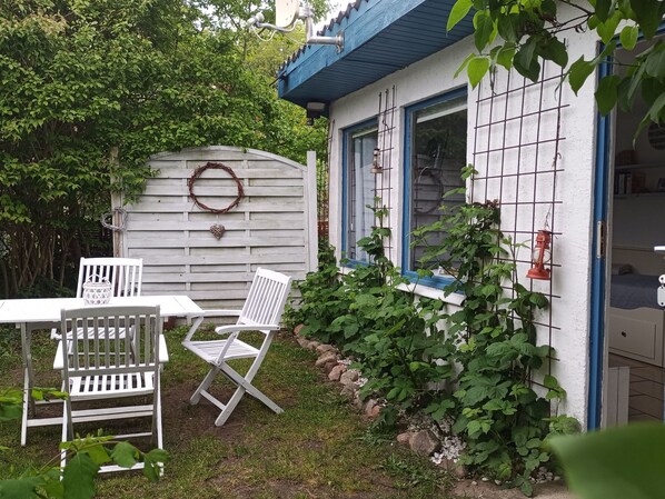 Una pequeña casa con fachada blanca y detalles azules. Tiene una mesa blanca y dos sillas blancas en el exterior.