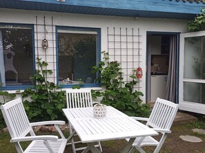 Un espacio exterior con mobiliario blanco, que incluye una mesa y dos sillas, una pared blanca con una ventana y una puerta.