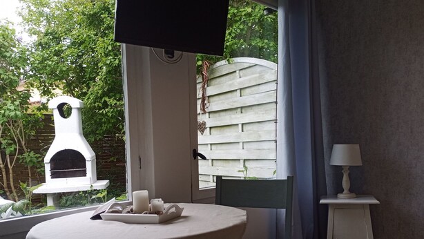 Une pièce avec une table et des chaises, un petit meuble blanc, un téléviseur fixé au mur et une vue sur la verdure à l’extérieur.