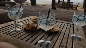 A wooden table with three glasses of Malfy Gin, a bowl of chips, and a plate of bread on a beach setting. - Calarossa-Red Island First Row House with Sea View and Double Terrace (Trinità d'Agultu e Vignola)