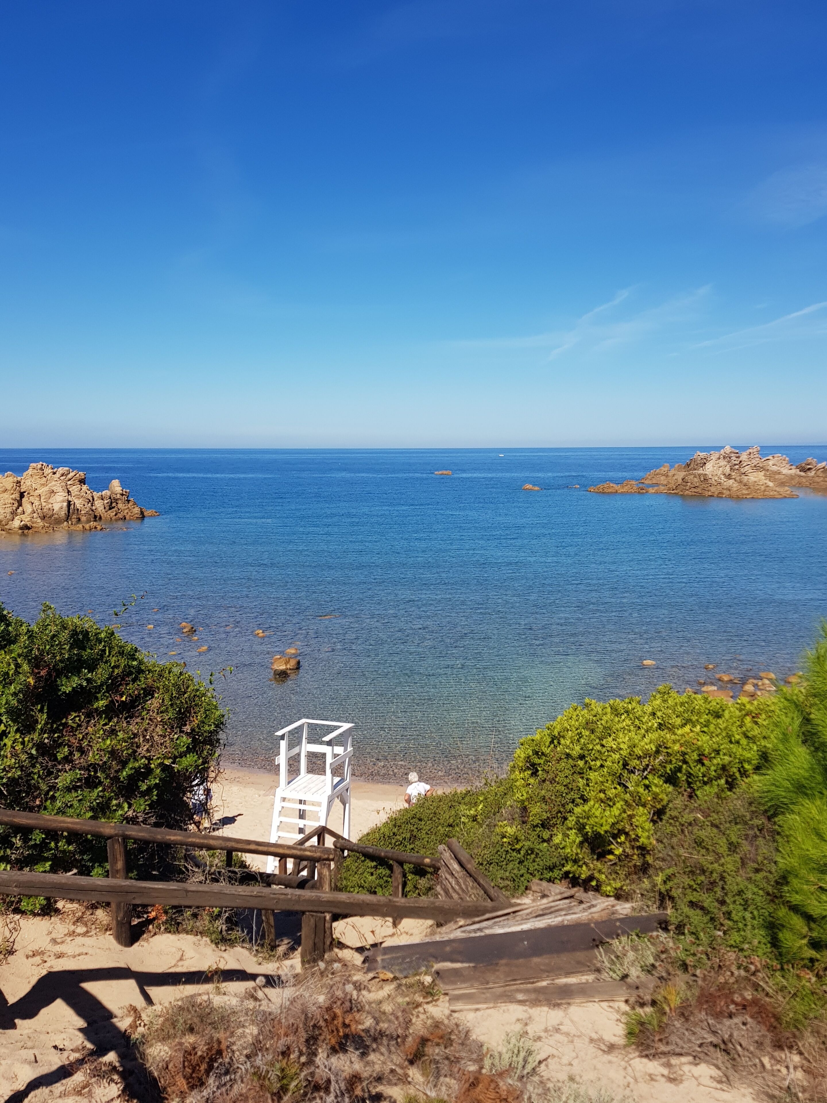 Blick auf die Küste mit einer Holztreppe, die zu einem Sandstrand führt, einem Rettungsturm und Felsformationen in der Ferne.