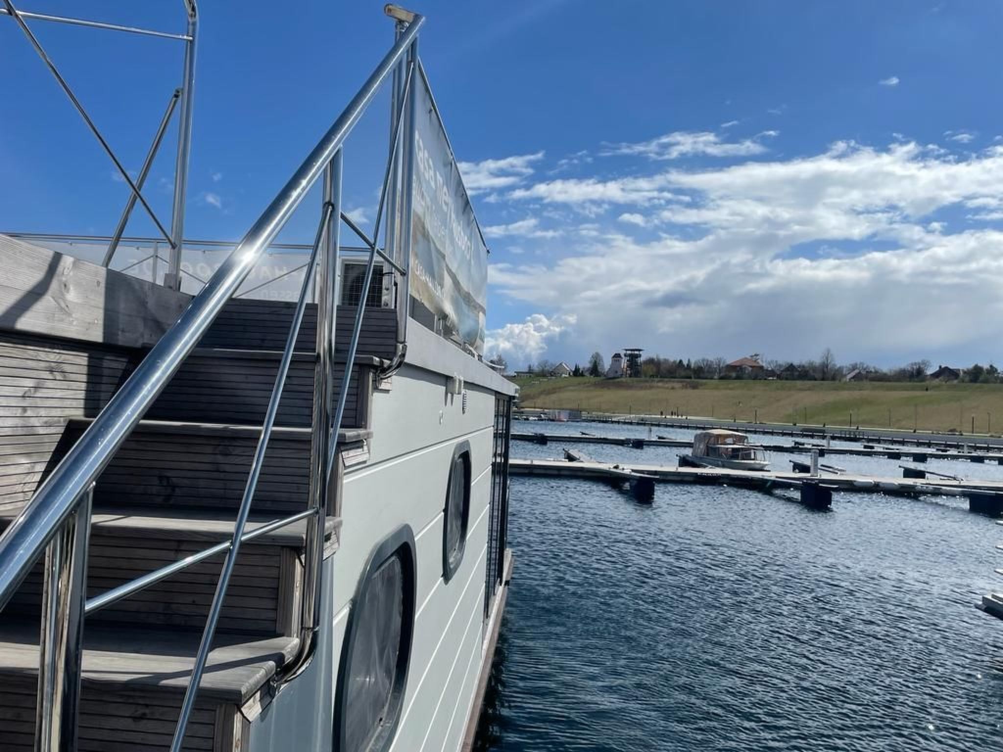 A boat with a wooden deck and metal railings, docked near a marina with calm water and a grassy landscape in the background.