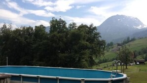 Piscina exterior con escalera, rodeada de césped verde y árboles, con una montaña al fondo.