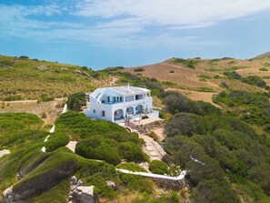 Una casa moderna blanca con techo plano, rodeada de exuberante vegetación y colinas.