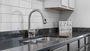 A kitchen with white tiled backsplash, granite countertop, stainless steel sink, and a sign with a message. - Ashley's Charming Chalet (Branson)