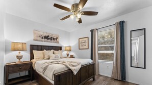 A bedroom with a wooden bed, two bedside tables with lamps, a ceiling fan, and a window with curtains. - Ashley's Charming Chalet (Branson)